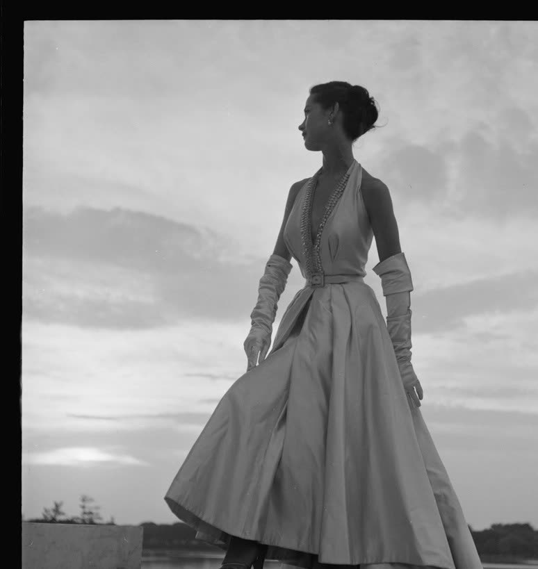Black and white photograph of a model in a halter evening gown and long gloves against an evening sky
